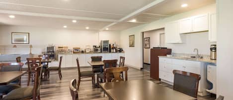 A spacious dining area with wooden tables and chairs, a kitchen with white cabinets, and a ceiling with recessed lighting.