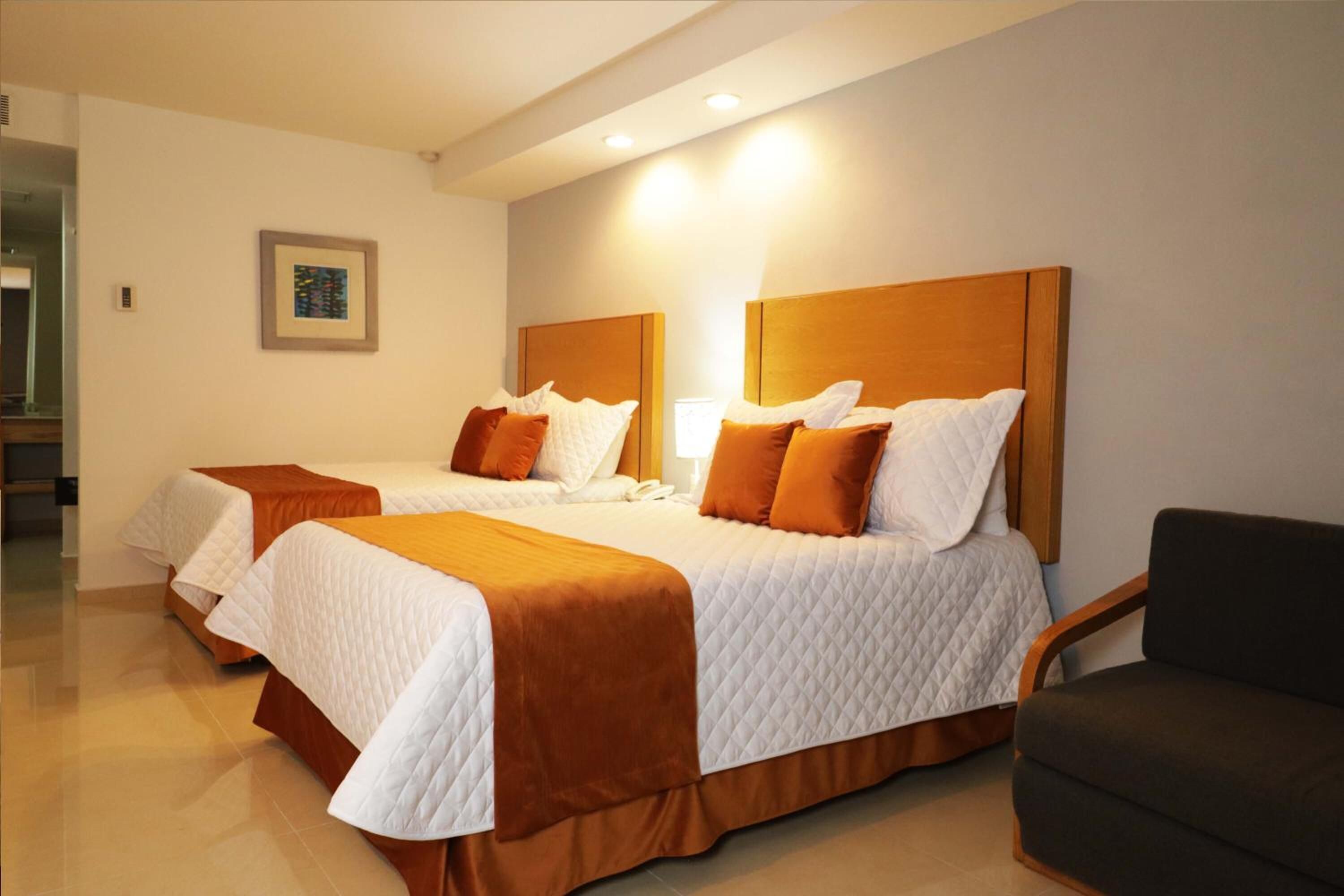 Habitación de hotel con dos camas, cada una con cabecero marrón, ropa de cama blanca y una manta marrón.