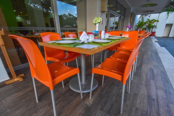 Un comedor con mesa de madera y sillas anaranjadas, un florero con flores y vistas al exterior a través de amplios ventanales.