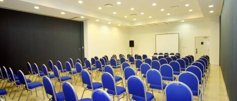 Un salón de conferencias con filas de sillas azules, un techo blanco con iluminación empotrada y una pantalla grande en la pared izquierda.