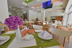 Un comedor con mesas dispuestas para el servicio, una pantalla grande instalada en el techo y un jarrón con flores moradas sobre la mesa.
