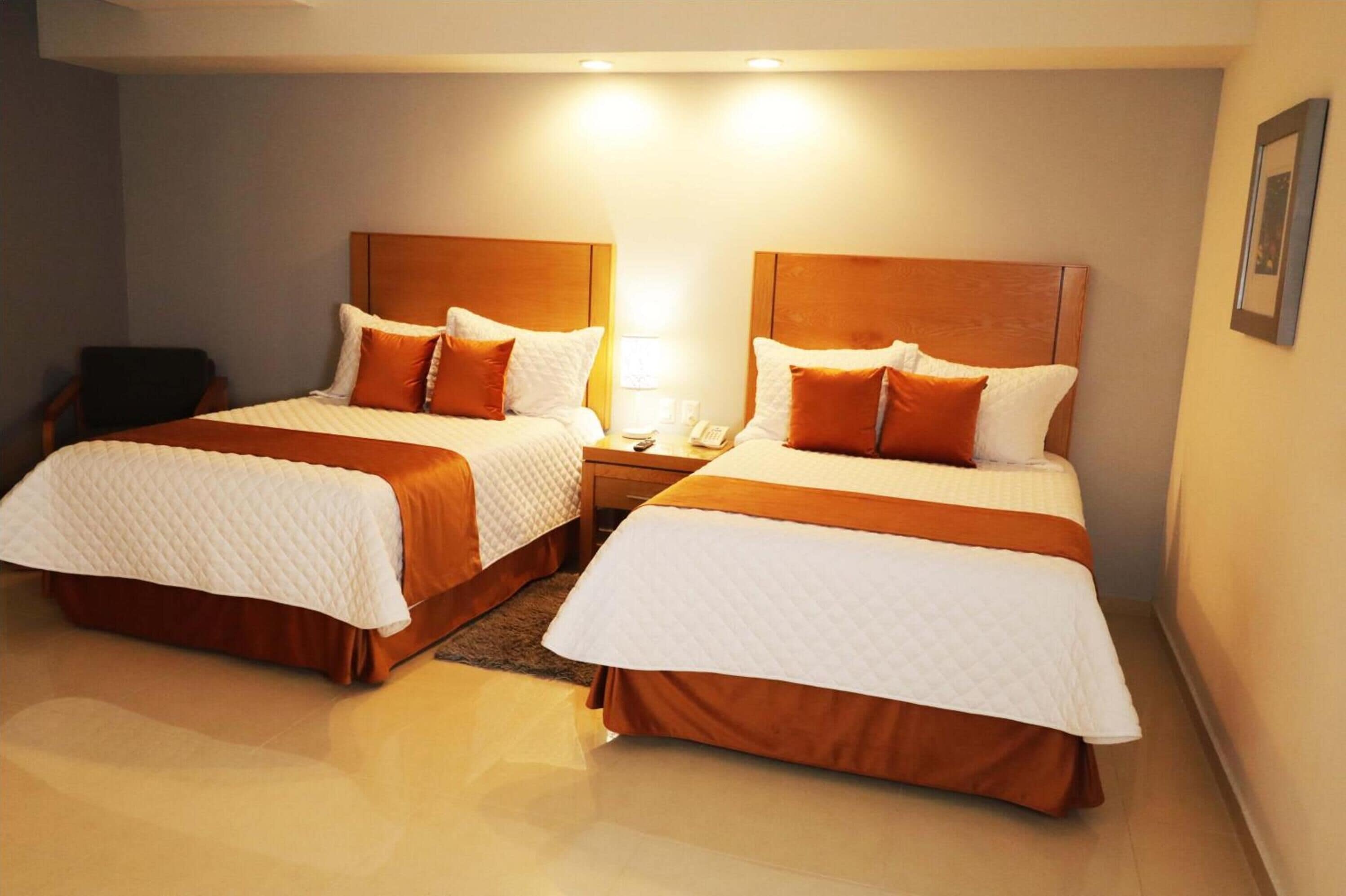 A hotel room with two beds, each with a wooden headboard, white bedding, and orange accents. There is a bedside table with a lamp and a phone.