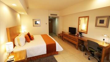 Quarto de hotel com cama, escrivaninha, cadeira, televisão e uma mesinha de cabeceira com abajur.