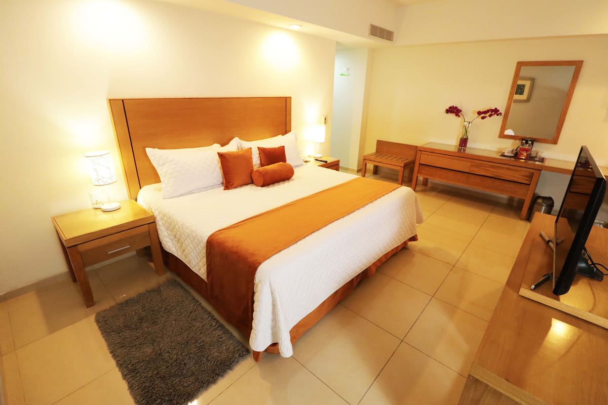 Ein Hotelzimmer mit einem großen Bett, einem Nachttisch aus Holz, einem Schreibtisch, einem Sessel, einem Fernseher und einem Spiegel.