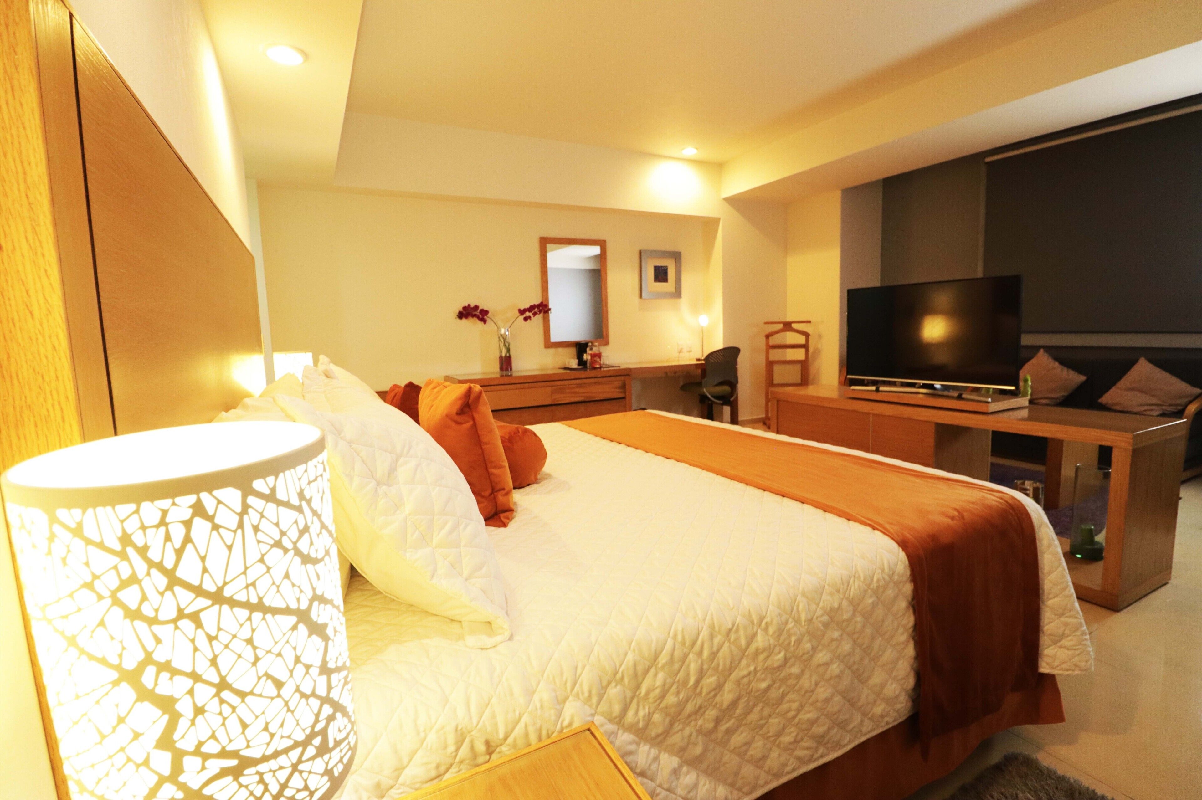A hotel room with a large bed, a desk with a flat-screen TV, a chair, and a small table with a vase of flowers.