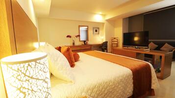 A hotel room with a large bed, a desk with a flat-screen TV, a chair, and a small table with a vase of flowers.