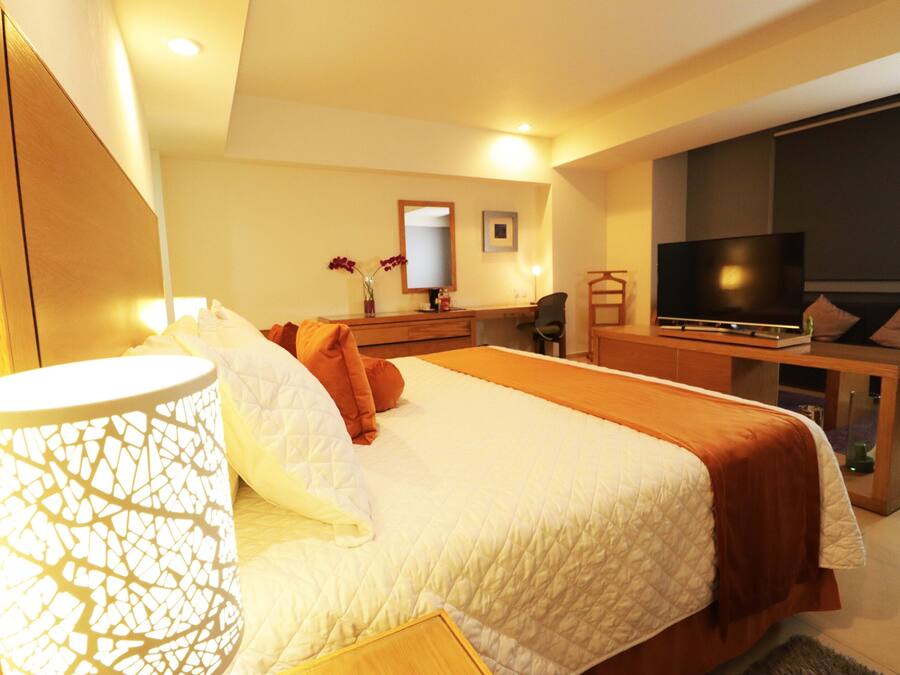 A hotel room with a large bed, a desk with a flat-screen TV, a chair, and a small table with a vase of flowers.