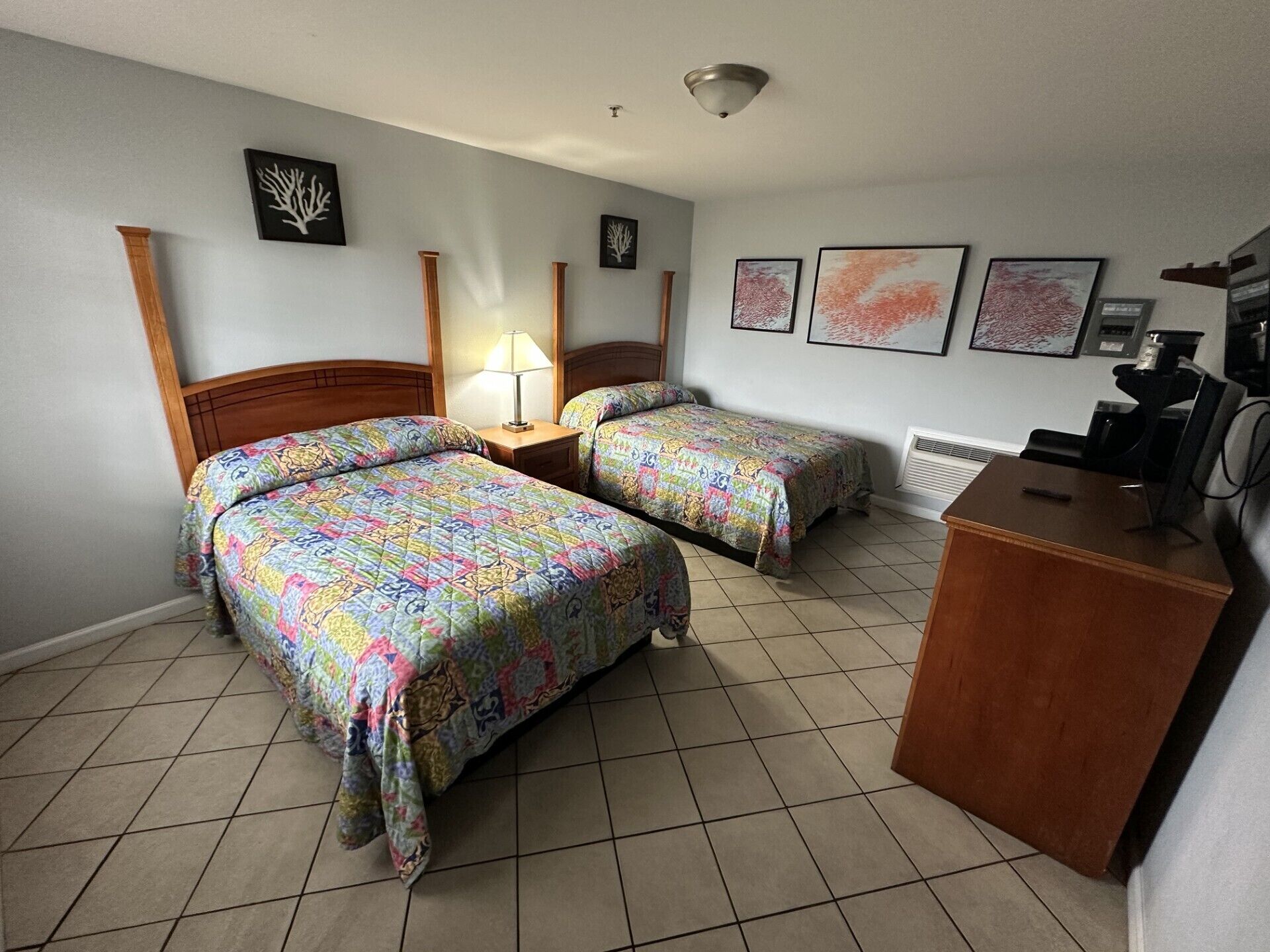 A hotel room with two beds, a desk, and a chair. There are wall-mounted artworks and a ceiling light.