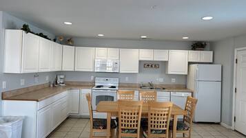 A modern kitchen with white cabinets, a stove, microwave, and a refrigerator. There is a dining table with chairs and a trash bin.