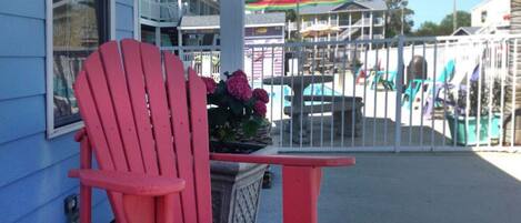 Une chaise Adirondack rouge sur un porche, avec vue sur un bâtiment et sur une zone délimitée par une clôture, où se trouvent des parasols colorés.
