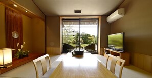 A dining area with a wooden table, chairs, a television, and a view of greenery outside.