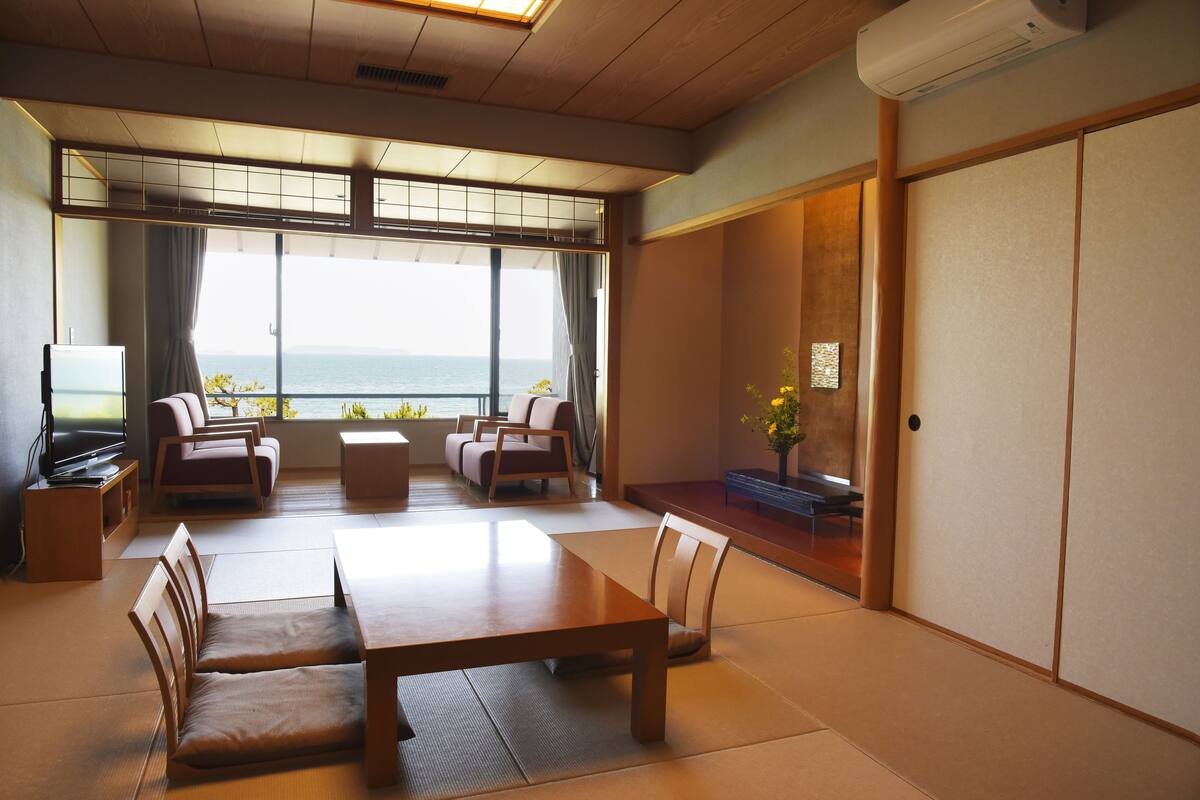 A traditional Japanese-style room with tatami flooring, a low table, and seating. A large window offers a view of the sea.