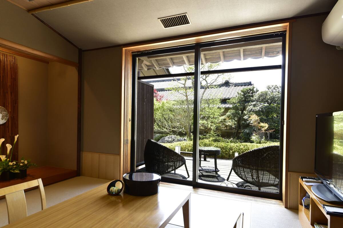 A room with a sliding glass door leading to a garden, a wooden dining table, a small black table, and a television on a wooden stand.