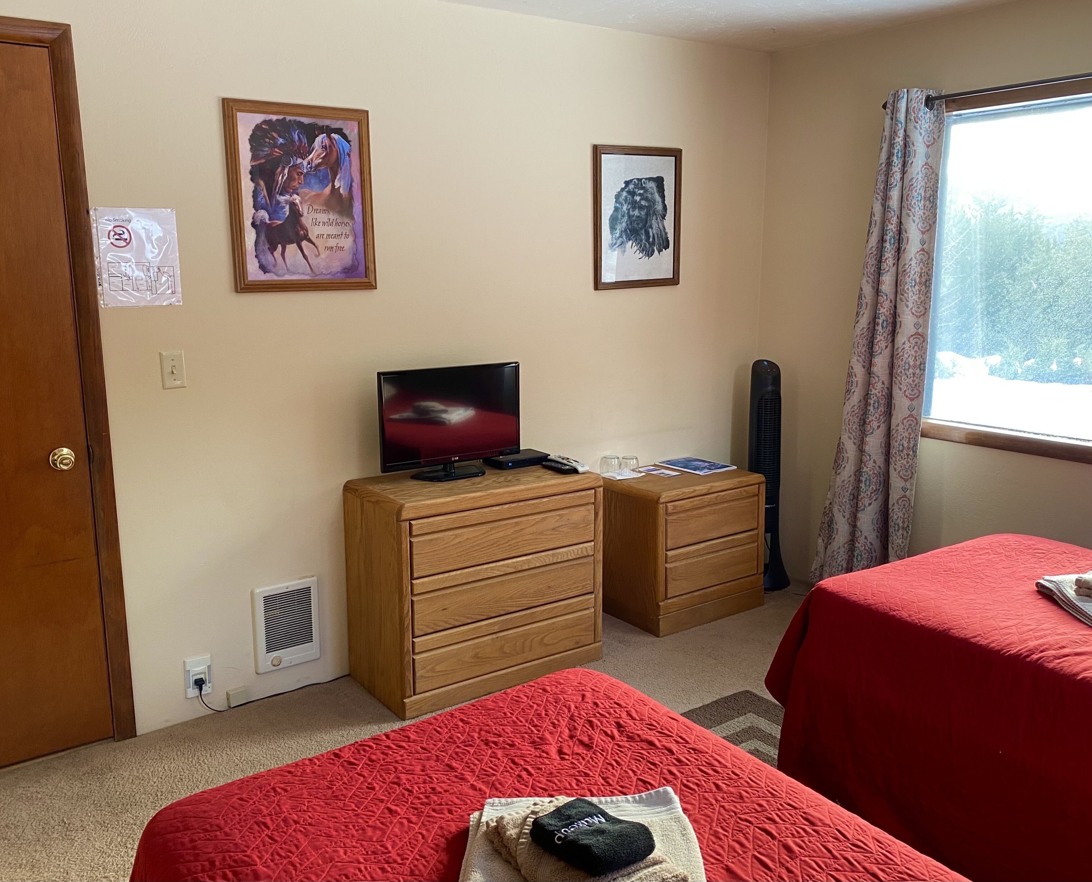 A hotel room with two beds, a TV on a wooden dresser, a window with curtains, and two framed pictures on the wall.