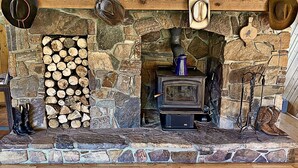 Una chimenea de piedra con estufa de leña, leños apilados a la izquierda y sombreros de vaquero colgados arriba.
