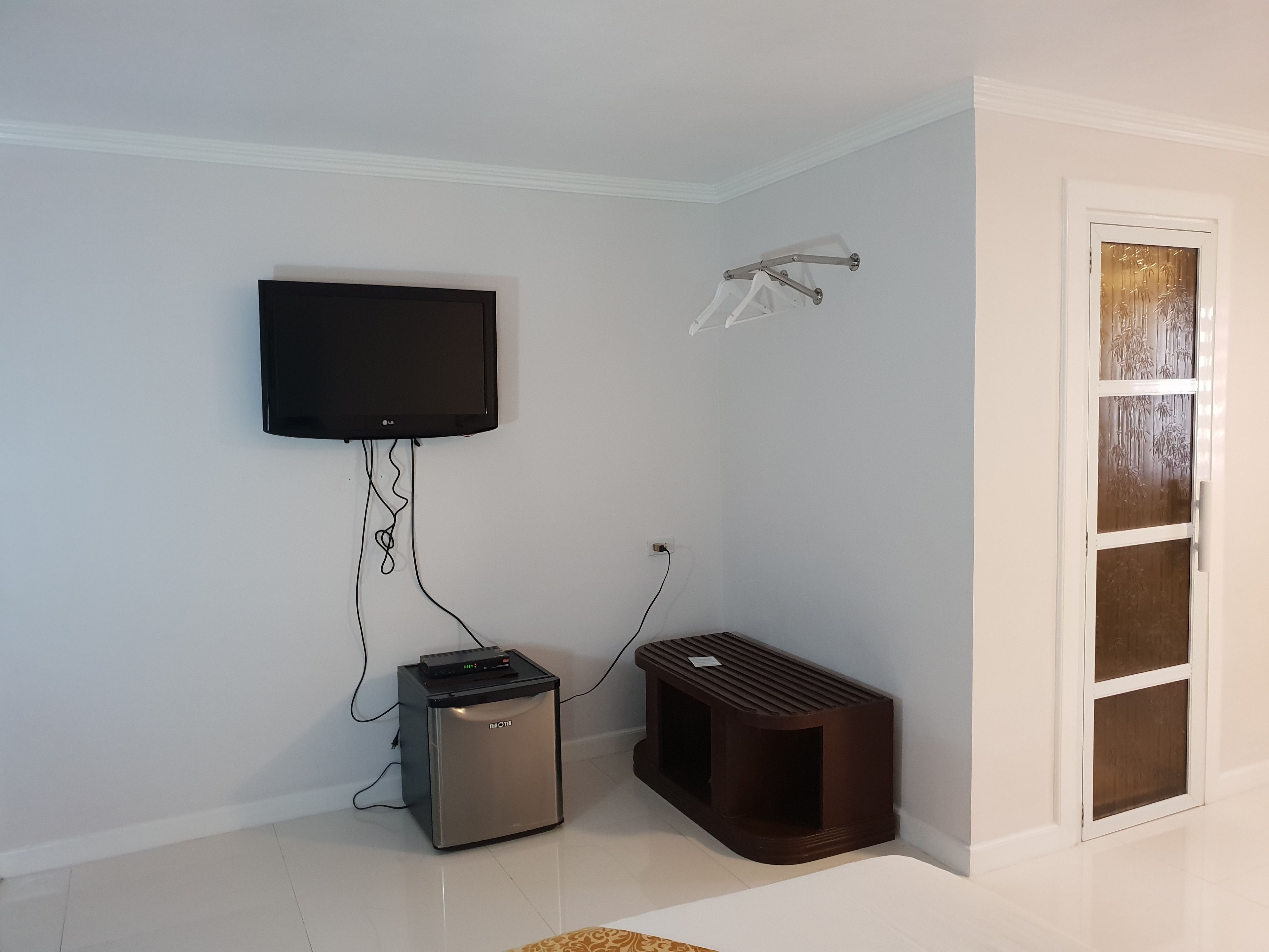 Una habitación con una televisión de pantalla plana montada en la pared, un refrigerador pequeño y un banco.