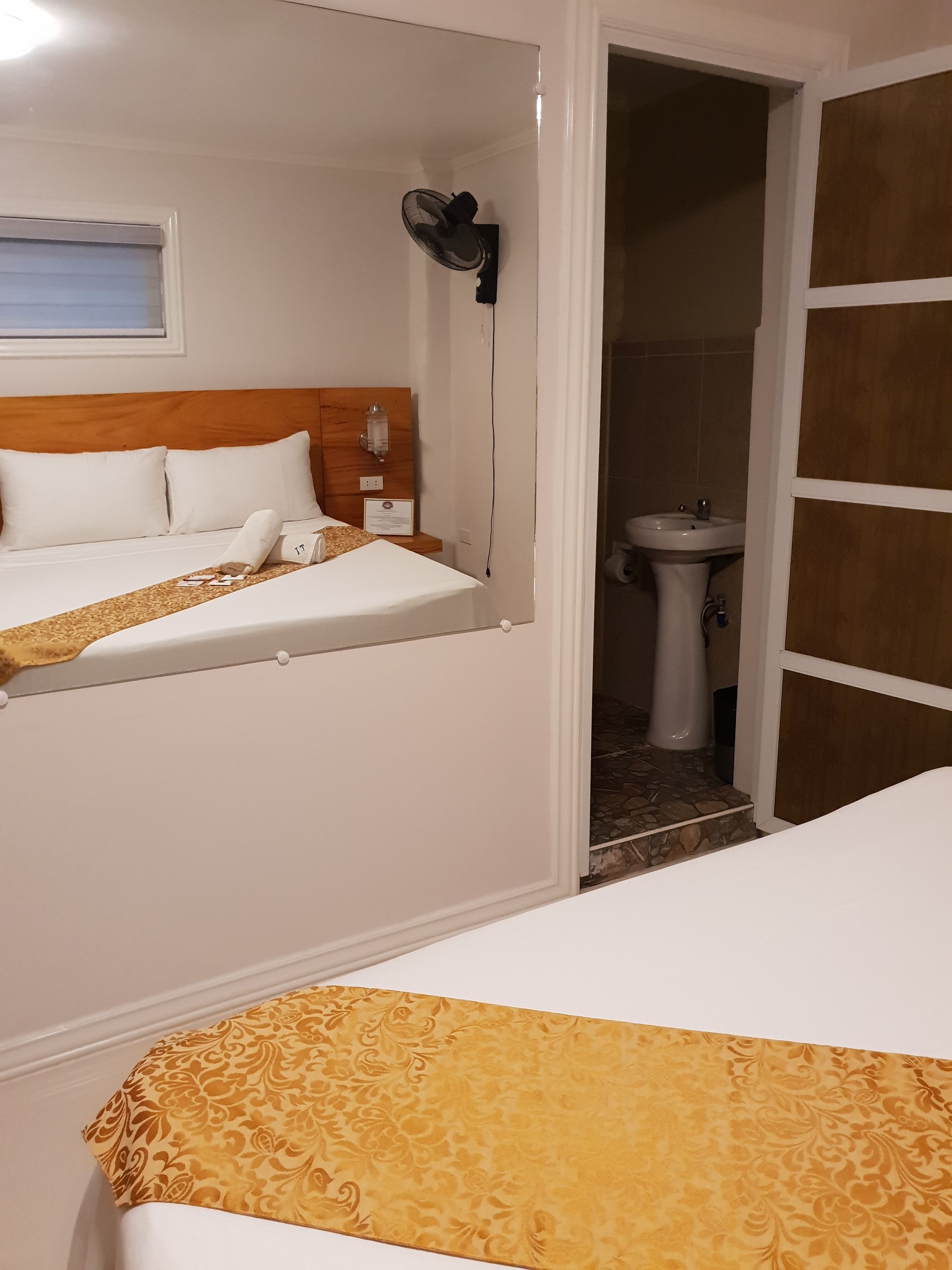 Ett hotellrum med två sängar, ett nattduksbord, en väggmonterad fläkt och ett badrum med handfat och toalett.