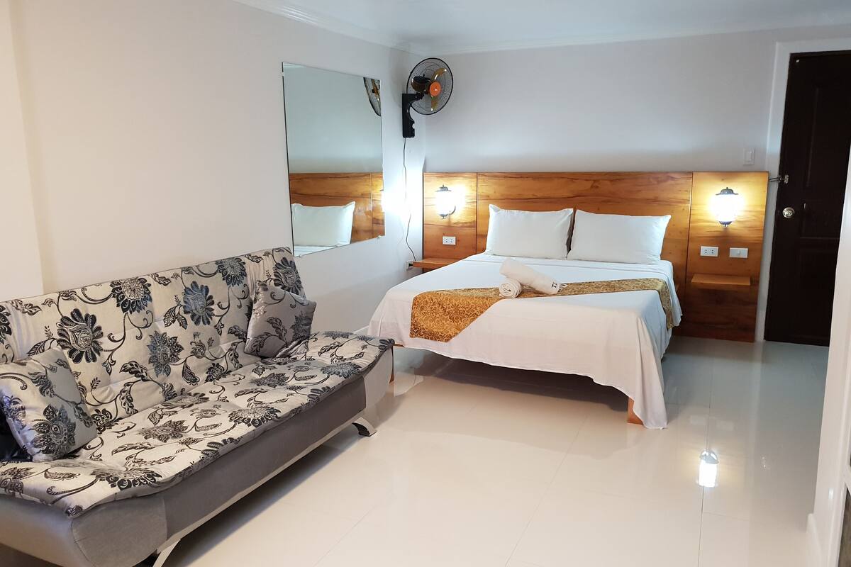 A hotel room with a bed, a sofa, a mirror, and wall-mounted lights.