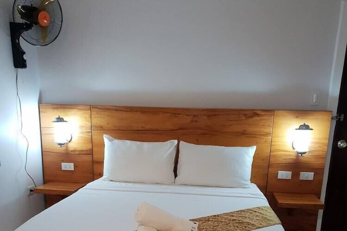 A hotel room with a wooden headboard, two bedside tables with wall-mounted lamps, a bed with white linens, and a rolled towel on the bed.