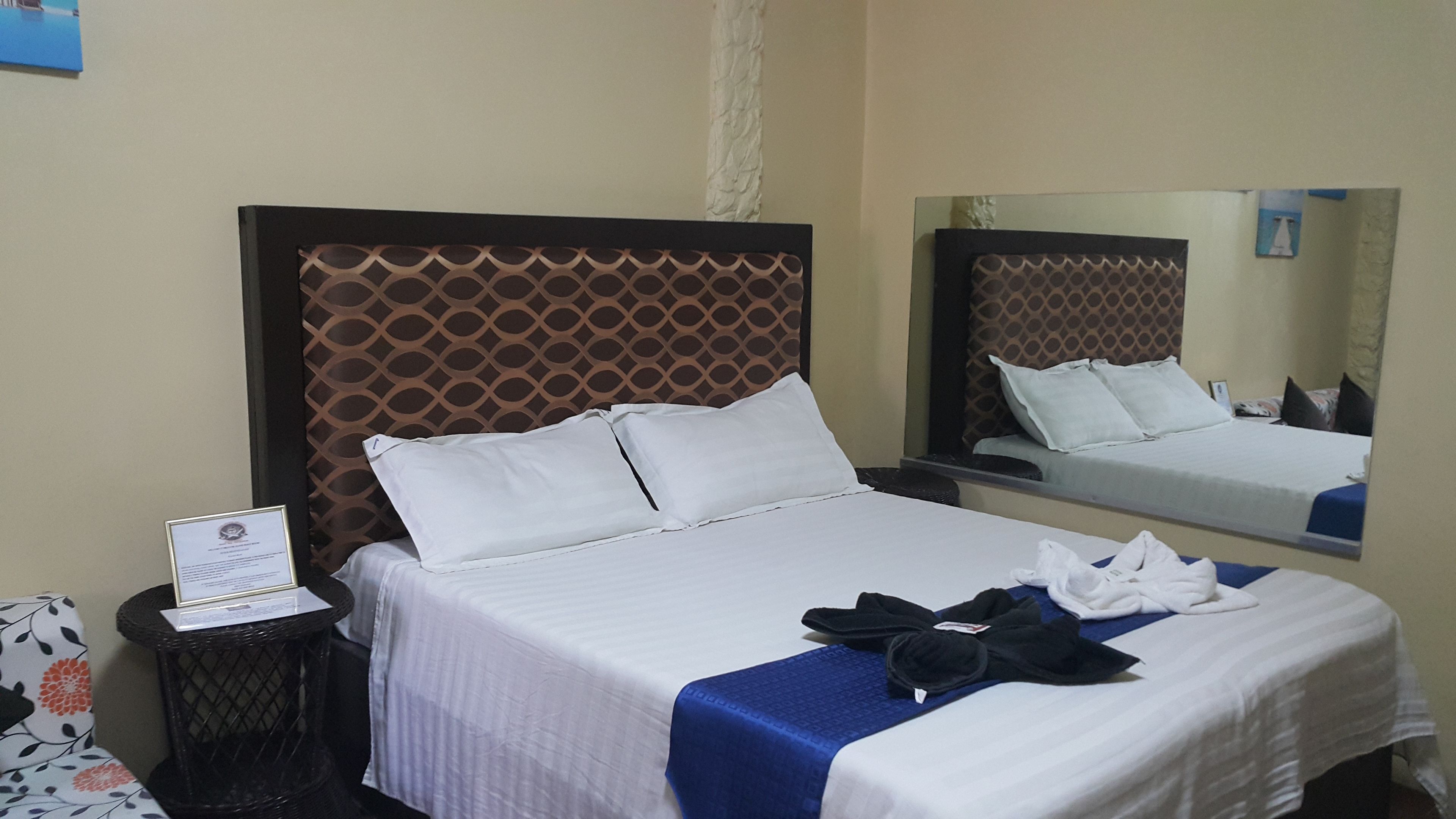 A hotel room with a large bed, a bedside table with a certificate, a small round table with a chair, and a mirror on the wall.