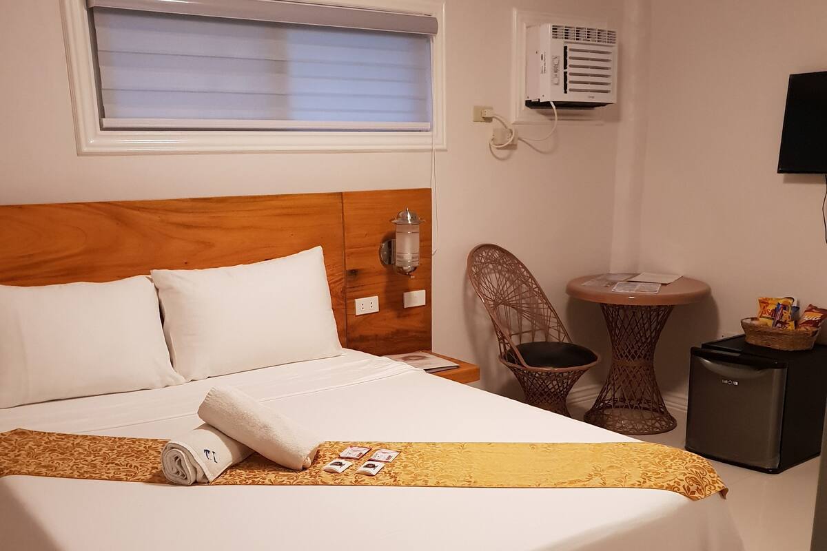 A hotel room with a large bed, a small table, a chair, an air conditioning unit, and a television.