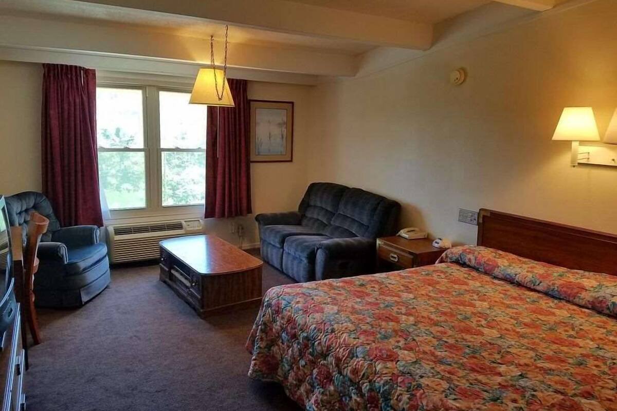 Une chambre d’hôtel comprenant un lit, deux fauteuils, une table basse en bois, un bureau avec un téléphone et une fenêtre avec des rideaux.