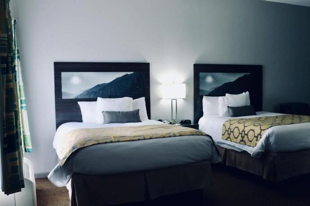 A hotel room with two beds, a nightstand, a lamp, and a view of mountains.