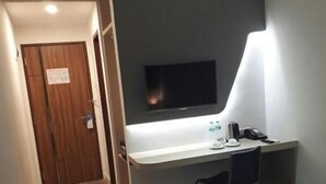 Ein kleines Zimmer mit Schreibtisch, Stuhl, Wand-TV und einer Lampe, die Licht spendet.