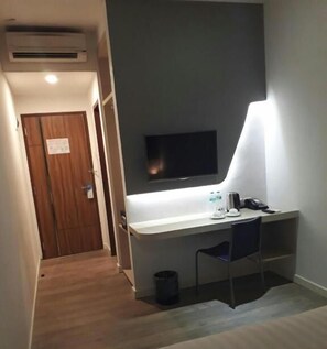 Ein kleines Zimmer mit Schreibtisch, Stuhl, Wand-TV und einer Lampe, die Licht spendet.
