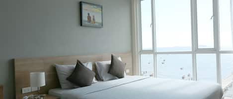 A hotel room with a large bed, a nightstand with a lamp, a painting on the wall, and a view of the sea through large windows.