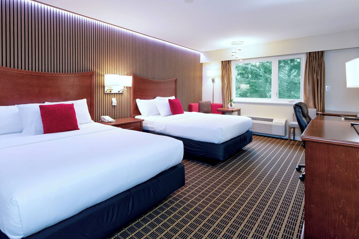 Ein Hotelzimmer mit zwei Betten, einem Schreibtisch, einem Stuhl, einem Fenster mit Blick auf Bäume und einer Wand mit vertikalen Holzleisten.