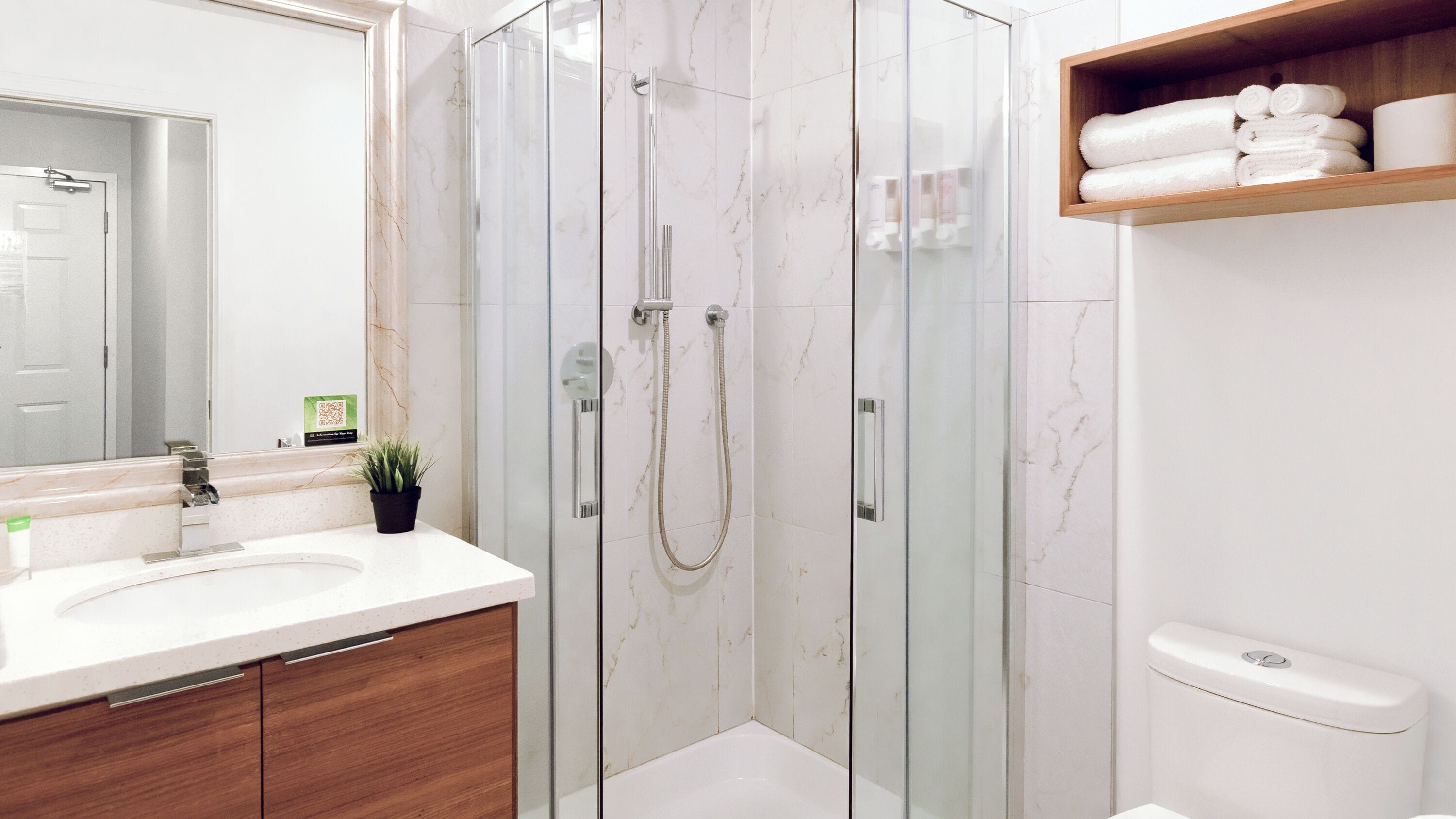 Une salle de bain moderne avec une douche à parois de verre, un lavabo en marbre blanc et une étagère en bois où sont posées des serviettes.