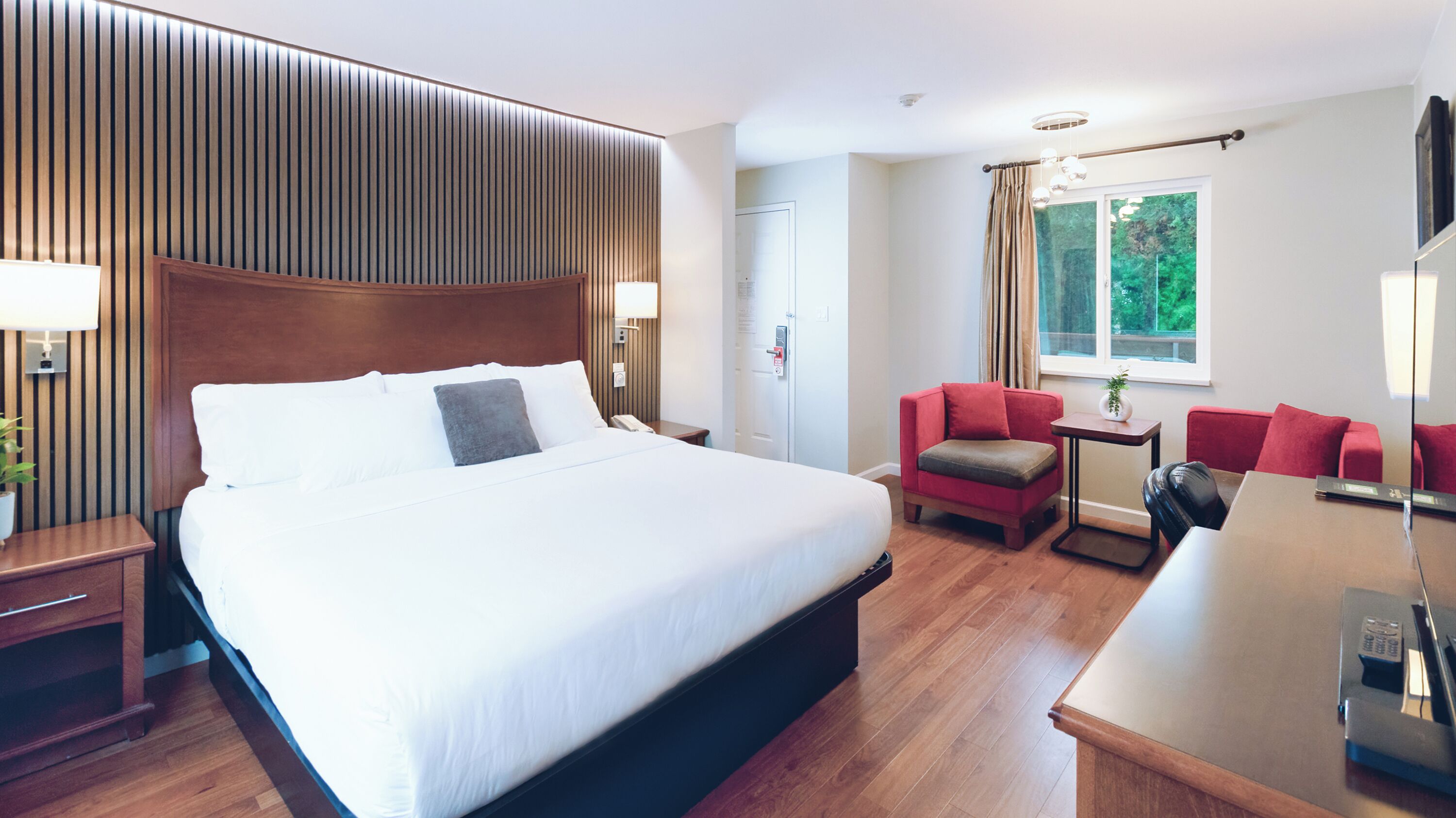A hotel room with a large bed, a desk with a chair, a red sofa, and a window with a view of greenery.