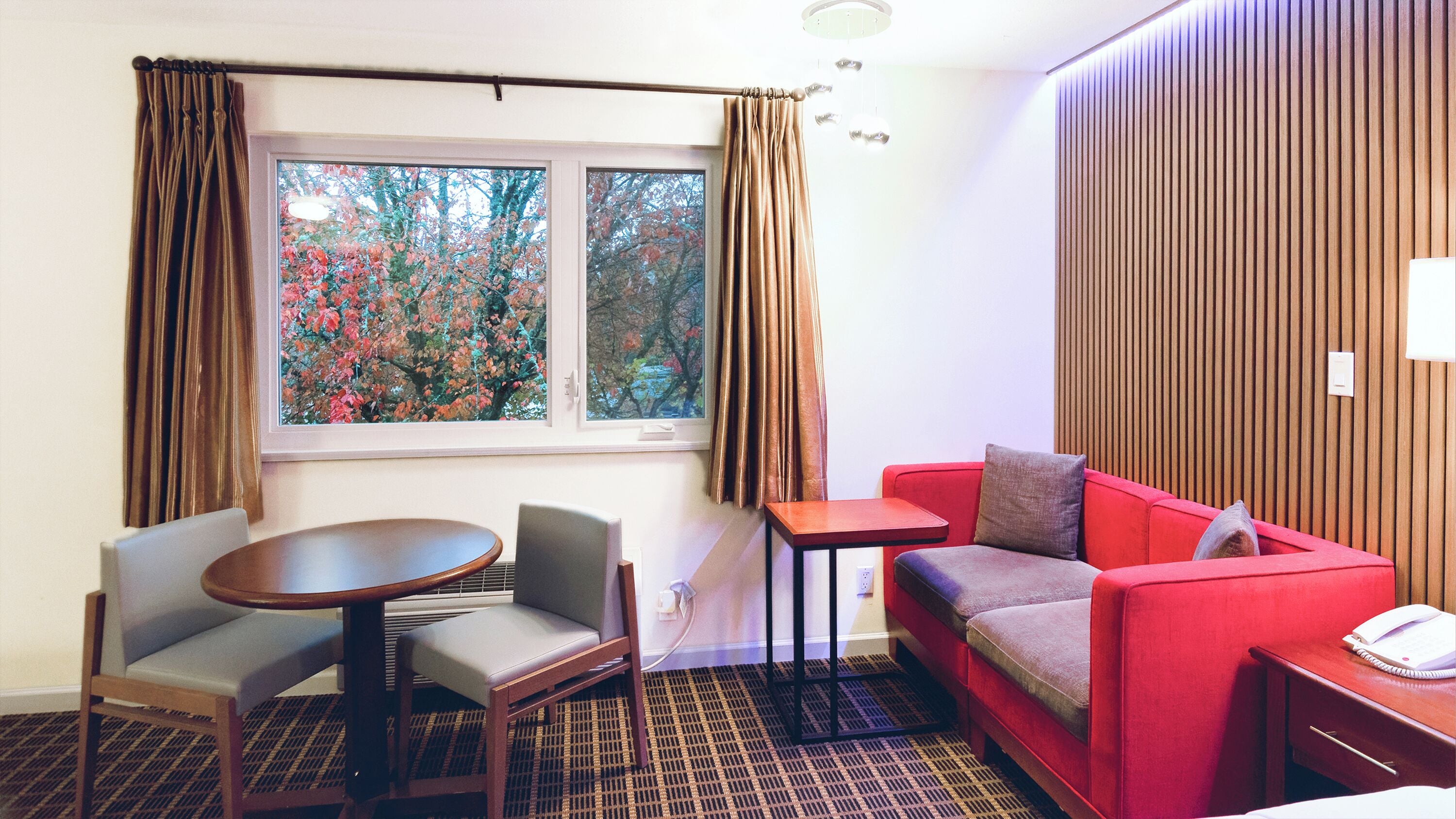 Een hotelkamer met een rode bank, een kleine tafel, een stoel, een raam met gordijnen en een houten wandpaneel.