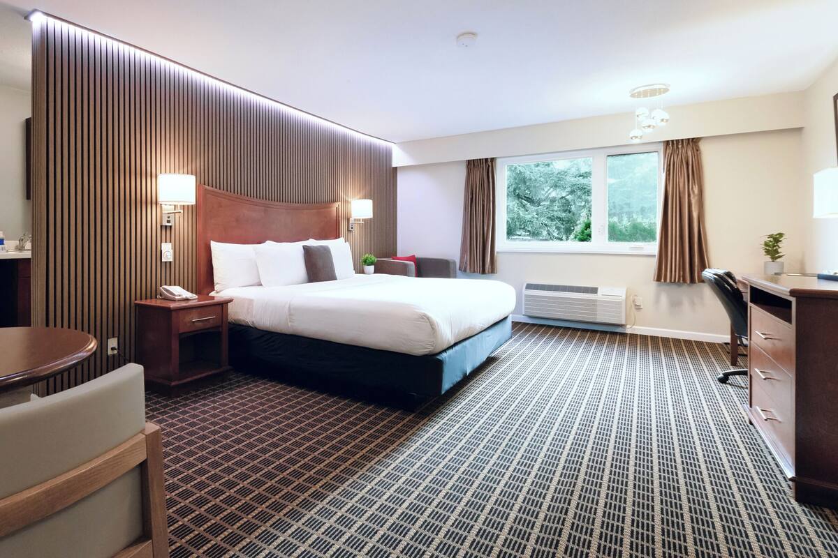 Ein Hotelzimmer mit einem großen Bett, einem Schreibtisch, einem Sessel, einem kleinen Tisch und einem Fenster mit Vorhängen.