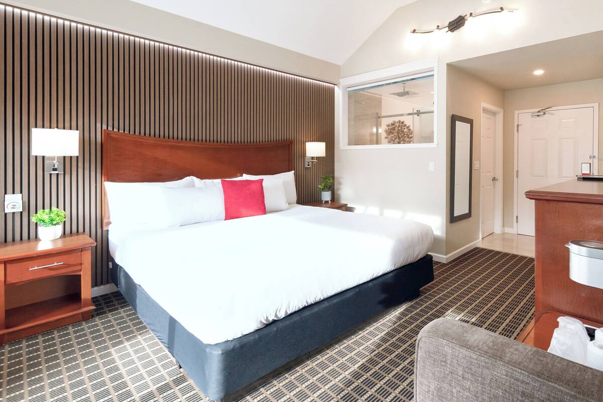 Ein Hotelzimmer mit einem großen Bett, einem Holz-Kopfteil, einem Nachttisch mit Lampe, einem Badezimmer mit Dusche sowie einem Schreibtisch mit Computer.