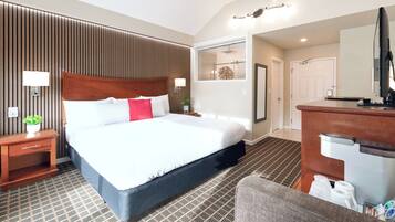 A hotel room with a large bed, a wooden headboard, a nightstand with a lamp, a bathroom with a shower, and a desk with a computer.