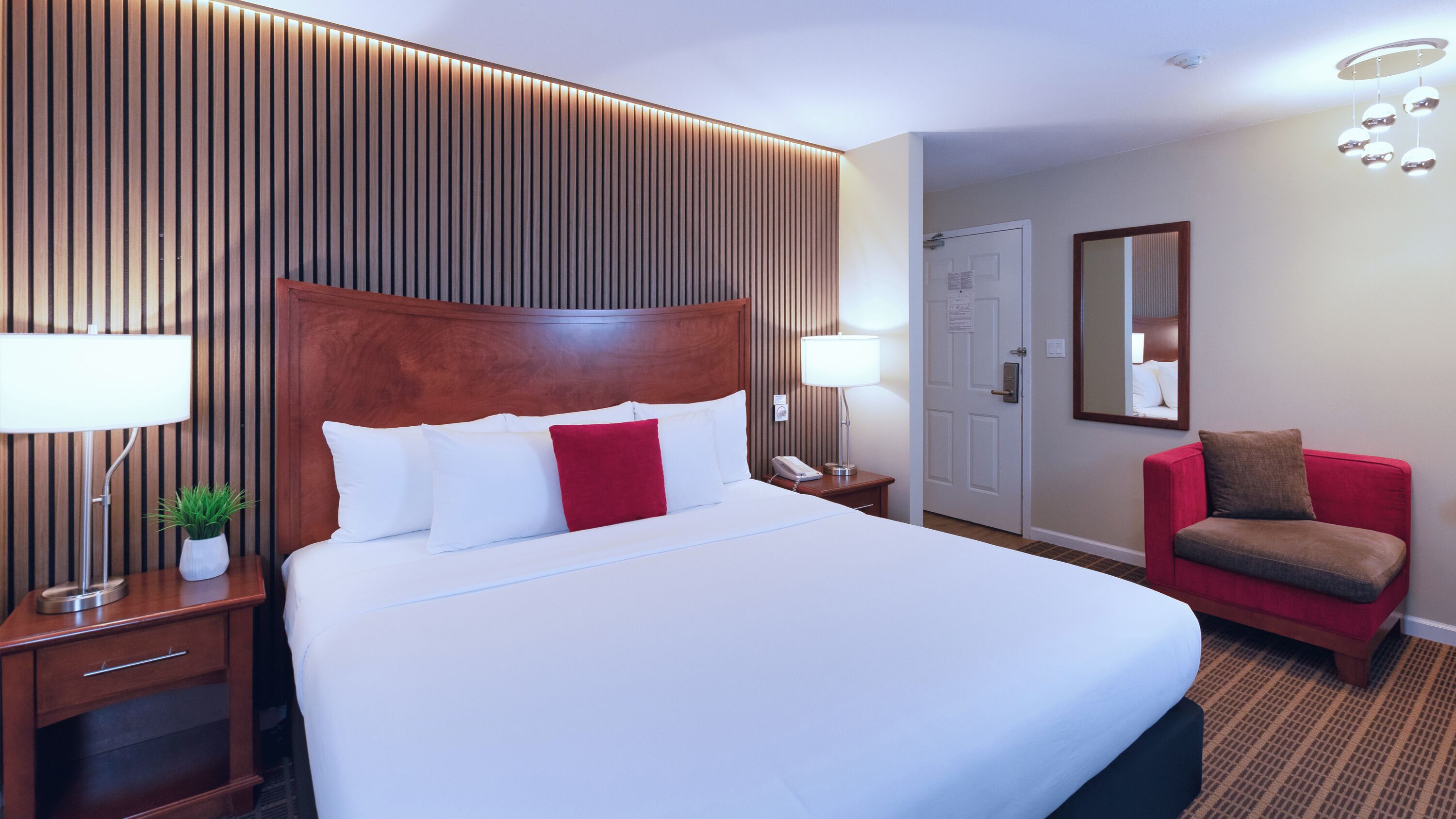 Ein Hotelzimmer mit einem großen Bett, Nachttischen, einem roten Sessel, einem Spiegel und einer Wandpaneele aus Holz.