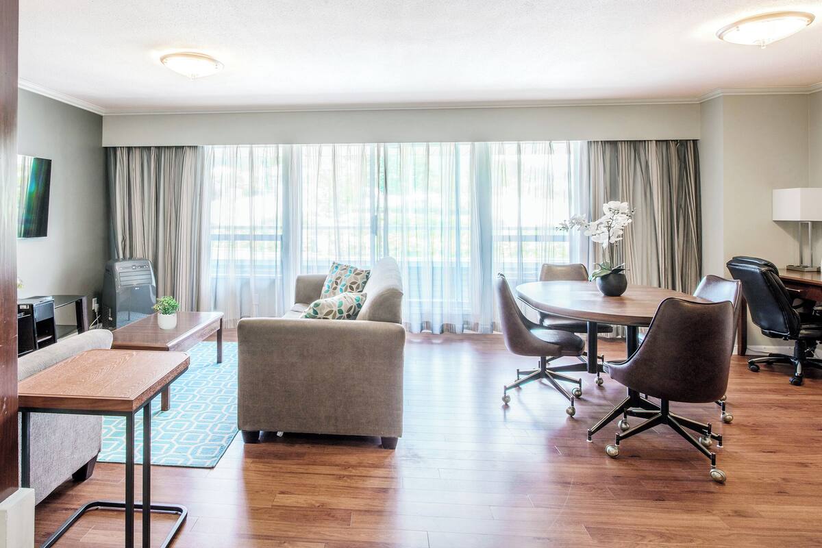 Ein Hotelzimmer mit einem Holztisch, einer Couch, einem Esstisch mit Stühlen, einem Fernseher und einem Fenster mit Vorhängen.