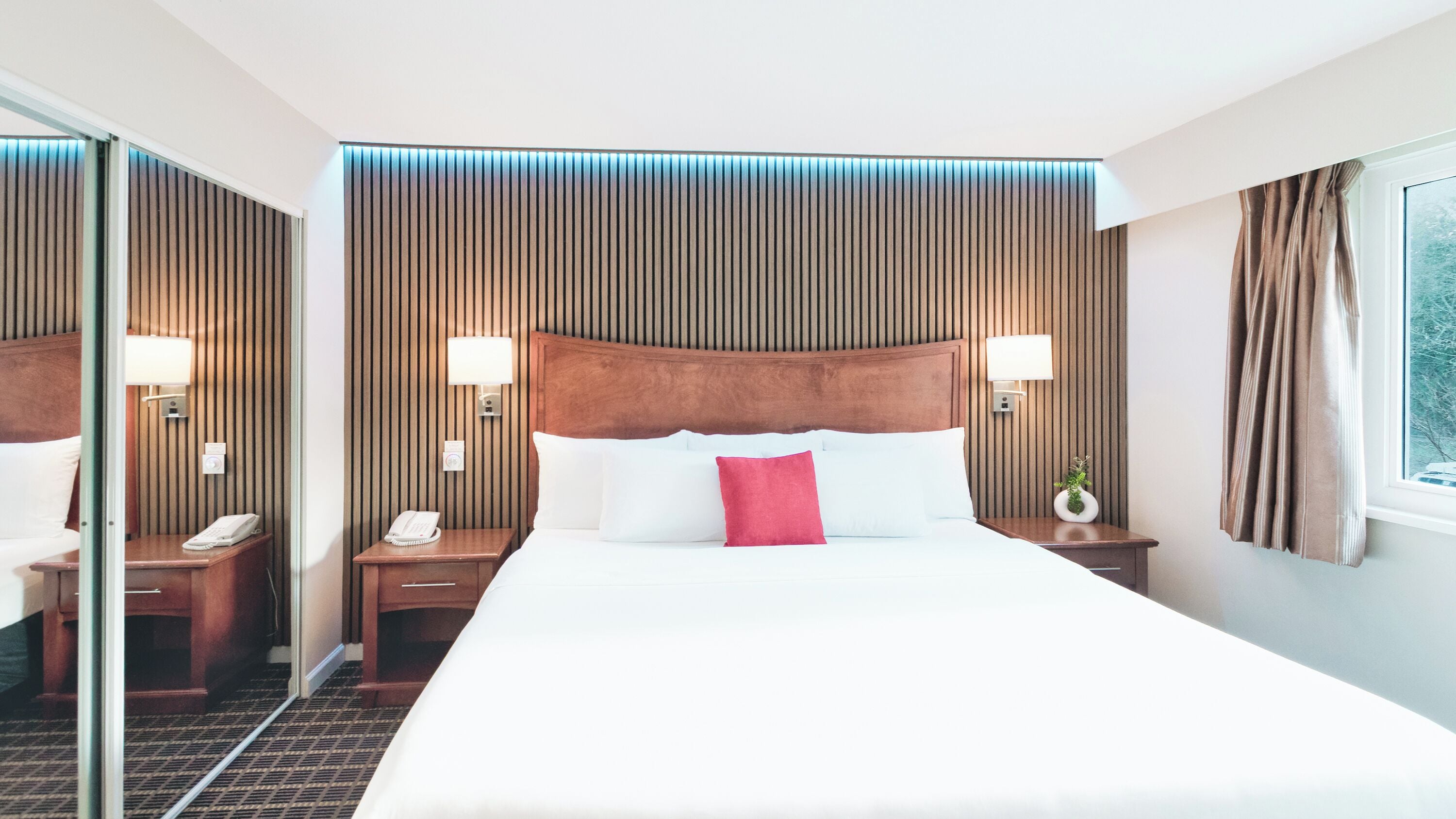 Habitación de hotel con una cama grande, cabecero de madera, lámparas de noche, teléfono y ventana con cortinas.