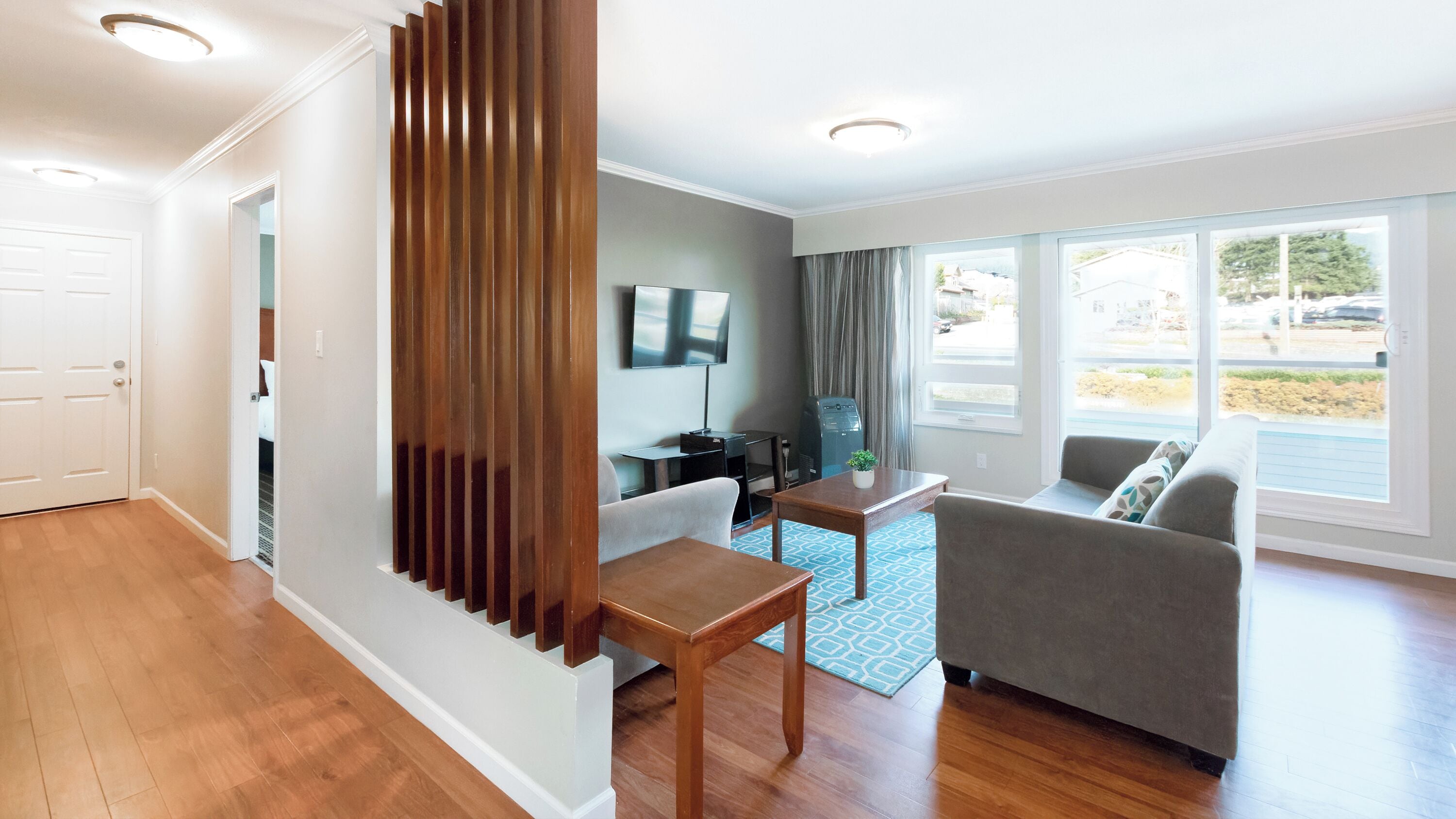 Un salón moderno con un separador de madera, un sofá gris, una mesa de centro de madera, un televisor y un ventanal con vistas al exterior.