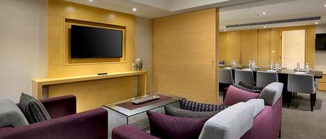 A modern hotel room with a flat-screen TV mounted on a wooden panel, a dining area with a table and chairs, and a seating area with a sofa and armchairs.