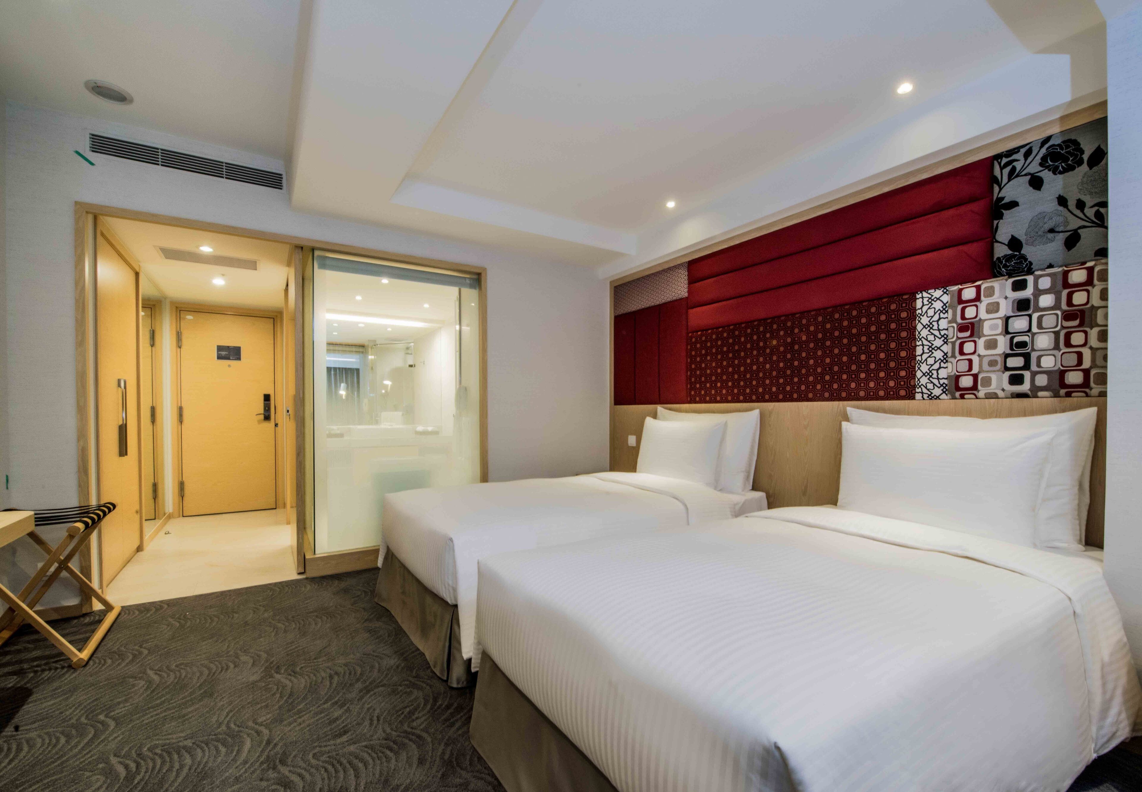 Habitación de hotel con dos camas, cabecero de madera, pared de acento roja y puerta de vidrio que da al baño.