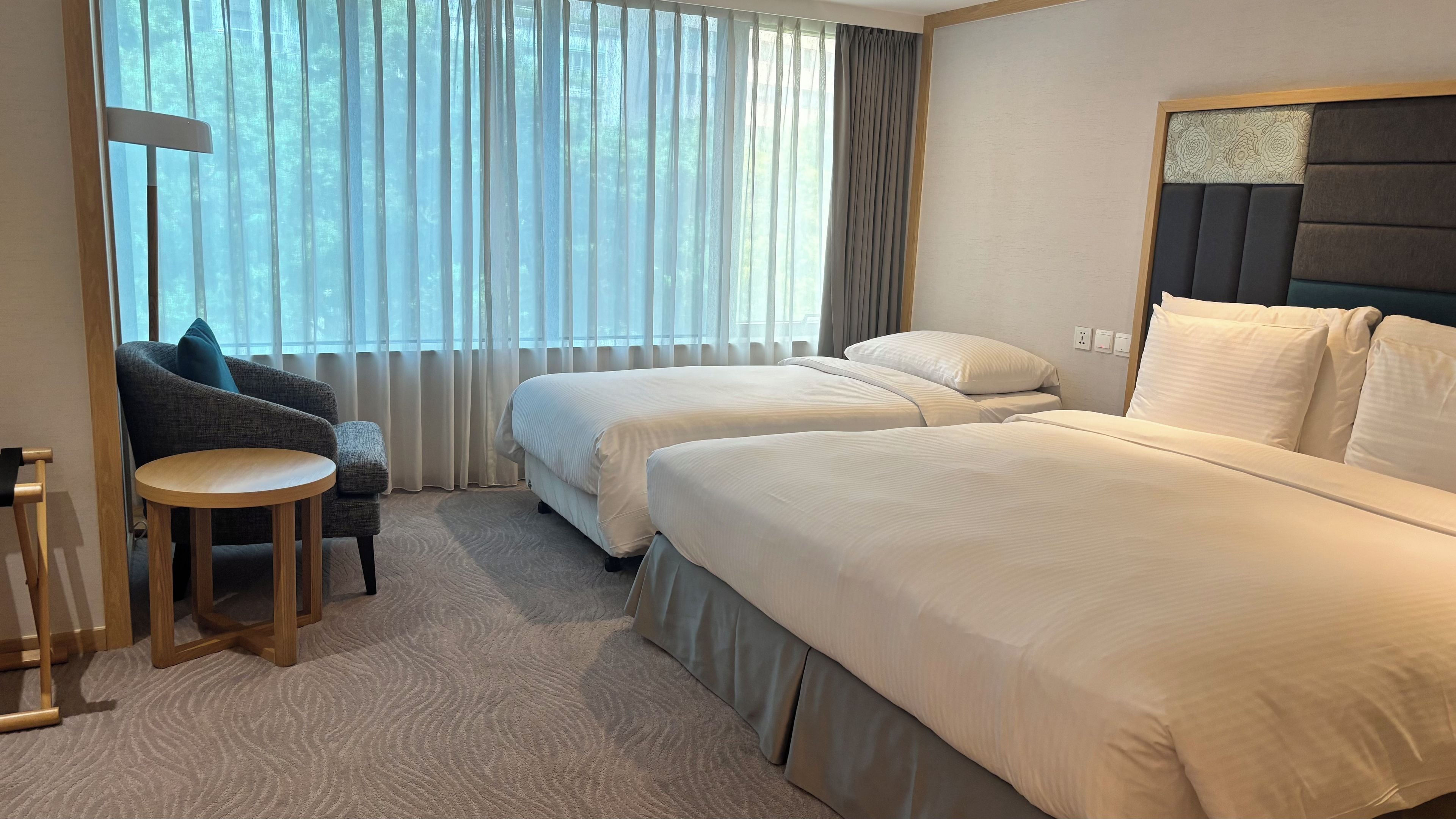 Habitación de hotel con dos camas, una silla, una mesita redonda y vistas a través de cortinas traslúcidas.