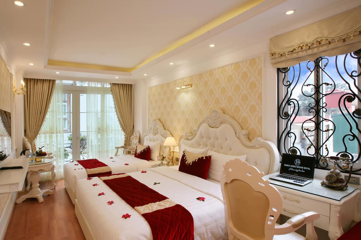 A hotel room with two beds, a desk, a chair, and a large window with decorative ironwork.