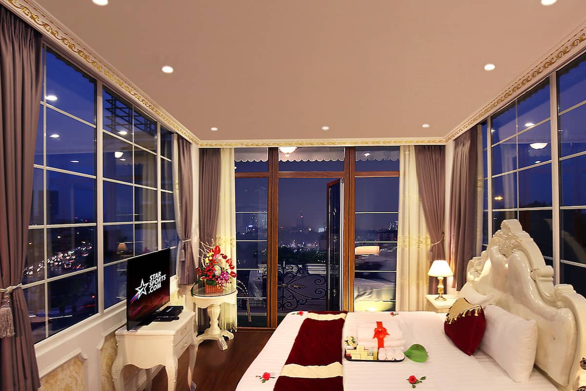 A hotel room with a large bed, a TV, a small table with a vase of flowers, and large windows with a city view.