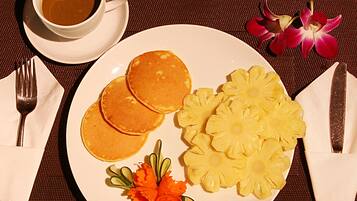 A plate with pancakes, pineapple slices, and a flower arrangement, accompanied by a cup of coffee and a flower on a table.
