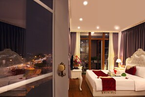 A hotel room with a large bed, a nightstand, a lamp, and a view of a cityscape through the window. - Hanoi Hotel Royal (Hanoi)