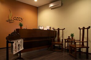 A reception area with a wooden reception desk, a potted plant, a chair, and a wall-mounted air conditioner. - COZY Boutique Guest House (Malang)
