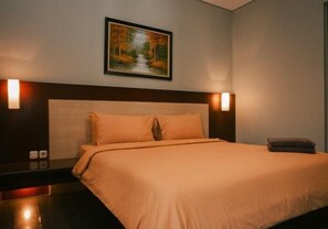 A hotel room with a large bed, a headboard, two bedside lamps, a nightstand, and a painting of a forest scene on the wall. - COZY Boutique Guest House (Malang)